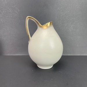 May include: A white ceramic jug with a rounded body and a curved handle. The rim and handle are accented with gold trim. The jug is likely for serving beverages and has a classic, elegant design.
