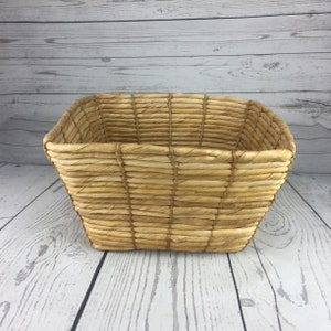 May include: A rectangular woven basket with a natural, light brown color. The basket has a slightly trapezoidal shape, with a wider opening and a narrower base. The weave pattern is horizontal and vertical, creating a textured surface.