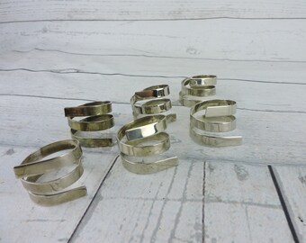 May include: A collection of silver-colored spiral napkin rings. Each ring is made of a flat, metallic band that coils around itself. The rings are arranged on a light-colored wooden surface, creating a simple, elegant display.