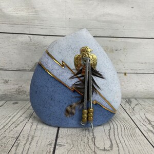 Könnte beinhalten: Eine dekorative, tropfenförmige Vase mit zweifarbigem Design. Die untere Hälfte ist ein gedämpftes Blau, während die obere Hälfte ein helles, gesprenkeltes Blau ist. Goldakzente und ein goldenes Adleremblem zieren die Vase, zusammen mit hängenden Federn und Perlen.