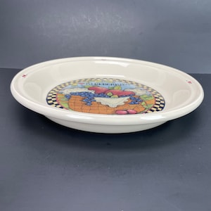 May include: Cream-coloured ceramic pie dish with a blueberry design. The centre shows a basket of blueberries, apples, and the word "BLUEBERRIES". The rim has small red heart accents. A charming addition to any kitchen.
