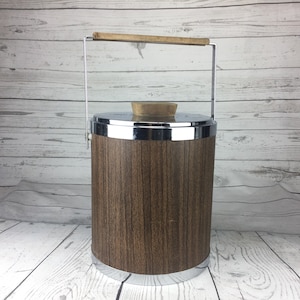 May include: A vintage ice bucket with a wood-grain finish and chrome accents. It features a stainless steel handle with a wooden grip and a lid with a cork stopper. The cylindrical bucket is approximately 25 cm tall.