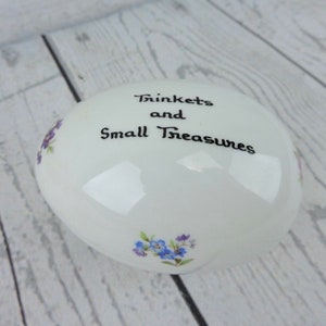 Puede incluir: Una caja de baratijas ovalada blanca con las palabras "Trinkets and Small Treasures" en escritura negra. Decorada con pequeños detalles florales azules y morados. Apoyada sobre una superficie de madera clara.