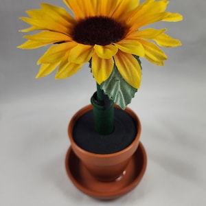 May include: A yellow sunflower with a brown center is attached to a green stem and placed in a brown terracotta pot with a saucer.