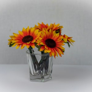 May include: A bouquet of artificial sunflowers in shades of yellow and orange, arranged in a clear glass vase. The sunflowers have dark brown centers and are set against a white background.