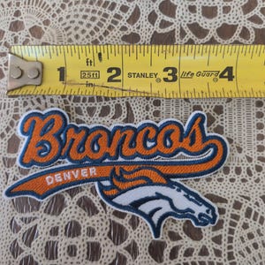May include: An embroidered Denver Broncos patch featuring orange and navy blue text and a white horse logo. A yellow Stanley measuring tape is positioned above the patch, showing it measures approximately 4 inches in total width.