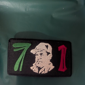 May include: Black rectangular patch with embroidered design. Features the number 7 in green, a portrait of a man in a hat, and the number 1 in red. The patch is likely for clothing or accessories.
