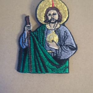 May include: Embroidered patch of a saint with a halo, holding a staff and a heart. The saint is wearing a green robe and a blue tunic. The background is a light brown.
