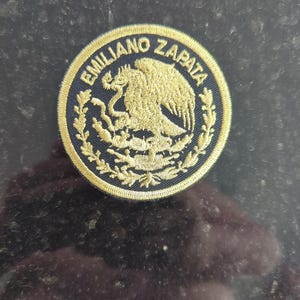 May include: Circular embroidered patch with the text "EMILIANO ZAPATA" in gold lettering around the edge. The patch features a gold eagle emblem in the center, surrounded by a dark blue border.