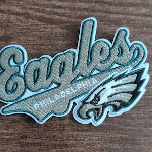 May include: An embroidered fabric patch featuring the word Eagles in a cursive font with the word Philadelphia printed below it. The design includes a stylized eagle head logo in shades of dark green, silver, and black with a white border.