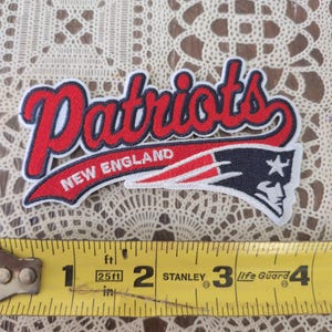 May include: An embroidered New England Patriots logo patch featuring red, white, and blue thread colors. The patch is positioned above a yellow Stanley measuring tape that shows the item is approximately 4 inches wide from end to end.