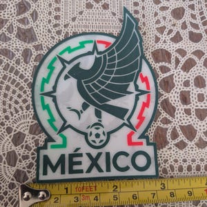 May include: A white, green, and red emblem featuring a dark green eagle with wings spread above a soccer ball, set within a circular frame. The word MÉXICO is printed in dark green below the emblem. A yellow measuring tape shows a width of 3 inches.