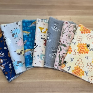 May include: A set of eight fabric squares with different patterns. The patterns include a blue and white pattern with the text "Save the Sea", a beige and brown pattern with a moose and bear, a gray pattern with the text "Coffee", a pink and green pattern with flowers and a bee, a yellow and orange pattern with honeycombs and a bee, a blue and white pattern with a sheep, a blue and white pattern with a turtle, and a blue and white pattern with a campfire and marshmallows.