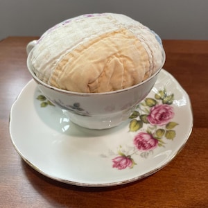 May include: A vintage teacup and saucer set with a floral design. The teacup holds a round, patchwork pincushion. The saucer and teacup are white with pink roses and green leaves. The teacup has a gold trim.