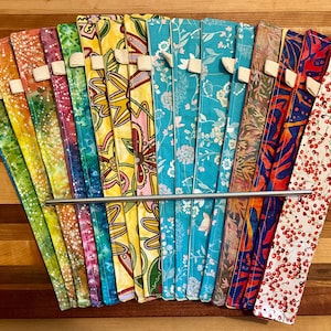 Puede incluir: Un conjunto de 15 pajitas de metal reutilizables envueltas en tela de colores. Las pajitas están dispuestas en forma de abanico y se mantienen unidas por una varilla de metal. La tela está estampada con varios diseños florales y geométricos.