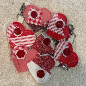 May include: A collection of handmade heart-shaped keychains in various red and white patterns. Each keychain features a large red button and a metal clasp. The fabrics include stripes, gingham, and solid colors, creating a charming, handcrafted aesthetic.