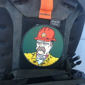May include: A green, round patch with a cartoon illustration of a man wearing a red hard hat with a flame logo, a yellow shirt with the text "Rough Rider" and a mustache. The patch is attached to a black backpack.