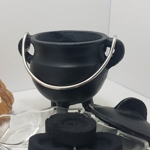 Puede incluir: Un caldero de hierro fundido negro con un asa plateada, una tapa y tres patas pequeñas. El caldero está junto a un trozo de calcita dorada en bruto y un plato de vidrio en forma de hoja con tres briquetas de carbón. Un tema de brujas.
