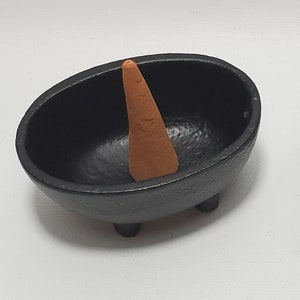 May include: Two black, oval-shaped incense holders with three small legs. One holder contains a brown incense cone. The holders have a textured surface and are set against a white background. These are ideal for burning incense.