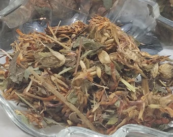 May include: Dried herbs in a clear glass dish. The herbs are a mix of green, brown, and reddish-purple. A clear plastic bag with a white label that reads "3 grams" is visible in the background. The herbs appear to be a natural product.