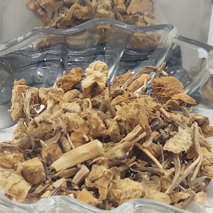 Puede incluir: Primer plano de hierbas secas de color marrón claro en un plato de vidrio transparente con forma de hoja. Dos pequeños paquetes de hierbas están en el fondo, uno etiquetado "3 gramos" y el otro con texto parcialmente oscurecido. Las hierbas parecen ser un producto natural.