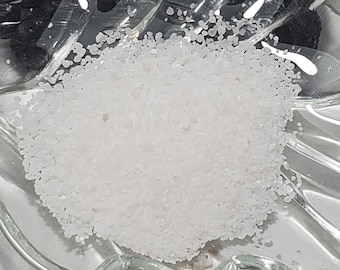 May include: A clear, leaf-shaped glass dish holds a pile of white, granular material and a section of dark, black material. The dish is on a light surface. A small label in the background reads "7 grams".