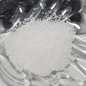 Puede incluir: Un plato de vidrio transparente en forma de hoja contiene un montón de material granular blanco y una sección de material negro oscuro. El plato está sobre una superficie clara. Una pequeña etiqueta en el fondo dice "7 gramos".