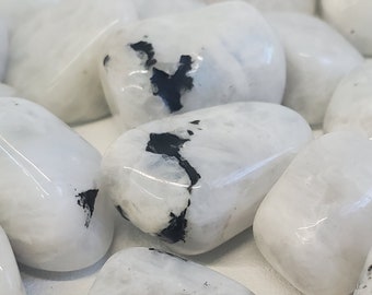 May include: A collection of polished, tumbled moonstones. The stones are primarily white with a pearlescent sheen, and some feature dark gray or black inclusions. The smooth, rounded stones are scattered across a surface.