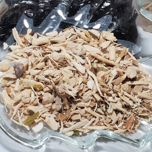 May include: A clear, leaf-shaped glass dish filled with light brown, dried herbs and spices. The dish is in front of a black, shiny substance and a small package with text. The herbs appear to be a mix of chopped roots and stems.