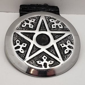 May include: A silver-coloured, circular pendant featuring a raised pentagram design. The pentagram is centred with a black circle and surrounded by decorative patterns. The pendant is likely made of metal and has a polished finish.