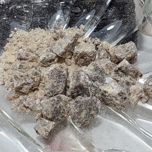 May include: A clear glass dish holds a pile of light brown, granular material and several dark brown, irregularly shaped chunks. The dish has a decorative, scalloped edge. The items appear to be some type of spice or food product.