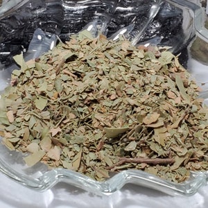 Op de afbeelding: Een heldere, blad-vormige glazen schaal gevuld met gedroogde, groene en bruine bladeren. De bladeren zijn klein en lijken een soort kruid of specerij te zijn. De schaal staat op een wit oppervlak.