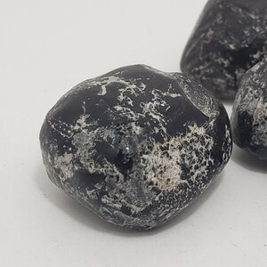 Puede incluir: Tres piedras negras pulidas con vetas blancas. Las piedras tienen una forma lisa y redondeada y un acabado brillante. Las piedras se exhiben sobre una superficie blanca. Estas piedras podrían usarse con fines decorativos o como elemento natural en manualidades.