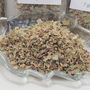 Puede incluir: Primer plano de hojas y flores de tilo secas en un plato de vidrio transparente. Las hierbas son una mezcla de verde, marrón y beige. Pequeñas etiquetas blancas con texto verde indican el peso de las hierbas en gramos.