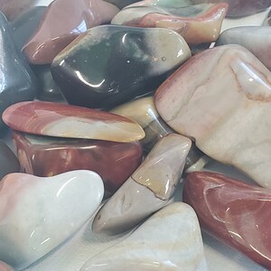 Op de afbeelding: Een verzameling gepolijste stenen in verschillende kleuren, waaronder rood, crème, grijs en groen. De stenen hebben gladde, afgeronde vormen en een glanzende afwerking. De afbeelding toont een close-up van de stenen, waarbij sommige elkaar overlappen.