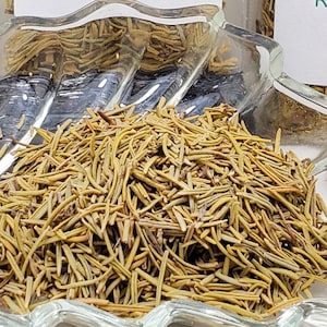 May include: Dried rosemary in a clear glass dish. The rosemary is a light brown colour and fills the dish. A label in the background reads "Rosemary 7 grams". The dish is on a light-colored surface.