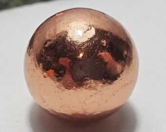 May include: A copper sphere with a hammered texture. The surface reflects light, creating highlights and shadows. The sphere is a solid, rounded shape, and the copper has a warm, reddish-brown hue. The object is likely a decorative item or component.