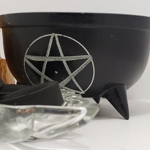 May include: A black cauldron with three legs and a white pentagram design. The cauldron is round and has a small rim. Incense and crystals are in the foreground, suggesting a spiritual or Wiccan theme.