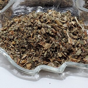 May include: A clear glass dish shaped like a maple leaf filled with a mound of dried, shredded brown and green herbal plant matter. The mixture consists of small, fragmented leaves, stems, and woody pieces, creating a textured, earthy appearance.