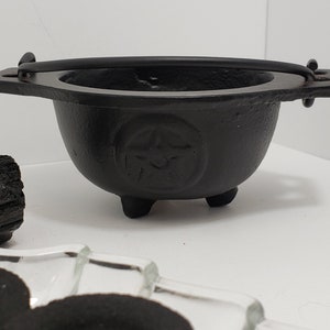May include: A small, black cast iron cauldron with a handle and three small feet. A pentagram is embossed on the side. The cauldron is likely used for rituals or decoration. A black crystal is next to the cauldron.