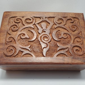 May include: A rectangular wooden box with a carved design on the lid. The box is a warm brown color, and the carved design features swirling patterns and a central stylized figure. The box appears to be handmade.
