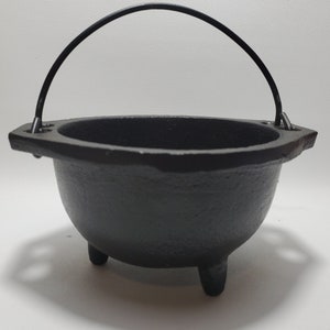 May include: A small, black cast iron cauldron with three short legs and a curved handle. The cauldron has a rounded body and a wide, flat rim. The handle is attached to the rim with metal loops. This item is suitable for decorative or functional use.