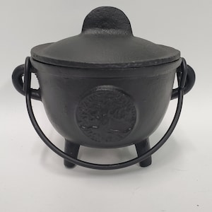 May include: A black cast iron cauldron with a matching lid, featuring a circular embossed tree design on the front. The pot has three short legs, two side handles, and a curved metal swing handle attached to the sides for carrying.