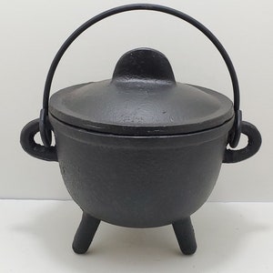 May include: A black cast iron cauldron with a rounded body, three short legs, and two side handles. It features a matching lid with a central handle and a curved metal bail handle attached to the sides for carrying or hanging.