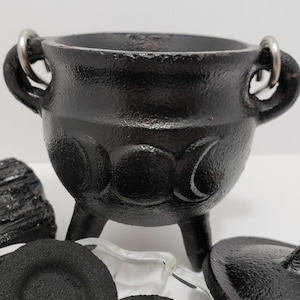 Puede incluir: Un caldero de hierro fundido negro con tres patas y dos asas, con un diseño de triple luna. El caldero está rodeado de discos de carbón vegetal, un trozo de turmalina negra y un plato de vidrio.