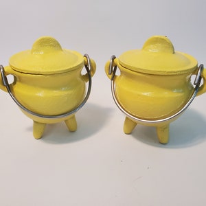 May include: Two small, bright yellow ceramic pots with lids and silver metal handles. Each pot has three small legs. The pots are likely decorative or for small items.