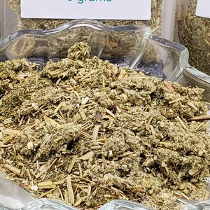 High-angle view of loose dried Mugwort (Artemisia vulgaris) displayed in a clear glass leaf-shaped ritual dish. The silver-green botanical texture is clearly visible against the white background, showcasing the quality of InSpyrit Metaphysical ritual herbs.