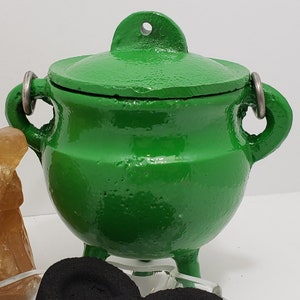 May include: A bright green, cast iron cauldron with a lid and three small feet. The cauldron has two metal handles and a loop on the lid. The cauldron is likely used for burning incense or other rituals.