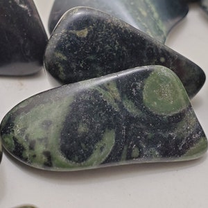 Puede incluir: Primer plano de varias piedras pulidas con una mezcla de colores verde oscuro y negro. Las piedras tienen una forma lisa y redondeada y un aspecto veteado, con patrones arremolinados de verde y negro. Las piedras probablemente se utilizan con fines decorativos o metafísicos.