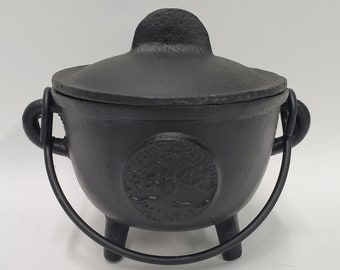 May include: A black cast iron cauldron with a matching lid, featuring a circular embossed tree design on the front. The pot has three short legs, two side handles, and a curved metal swing handle attached to the sides for carrying.